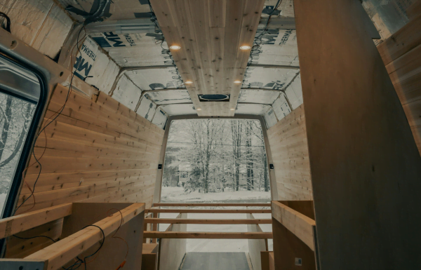 Interior view of a van in the process of being converted into a camper featuring wooden panels installed on the walls and ceiling visible wiring and a large rear window showing a snowy landscape with trees