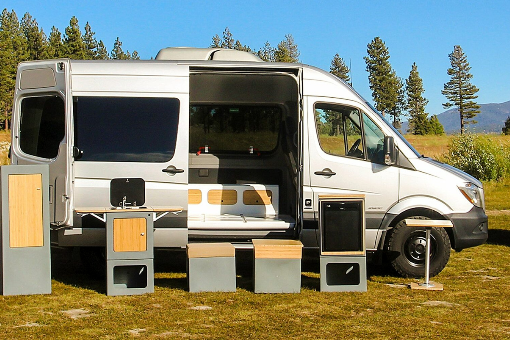 White camper van with its sliding door open, showcasing a modular outdoor kitchen setup and storage units on a grassy field with trees in the background.