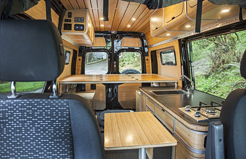 Modern camper van interior with kitchen, dining table, and wooden storage.