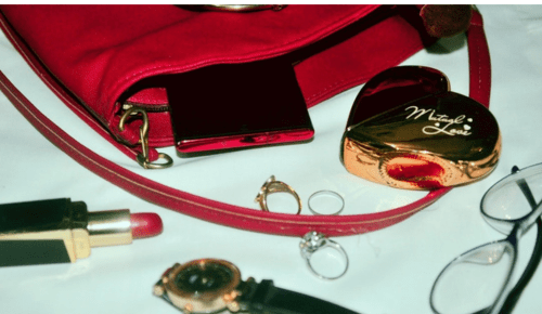 Sac à main rouge ouvert contenant des accessoires féminins, avec rouge à lèvres, montre, bagues, lunettes et un étui de maquillage posés autour.