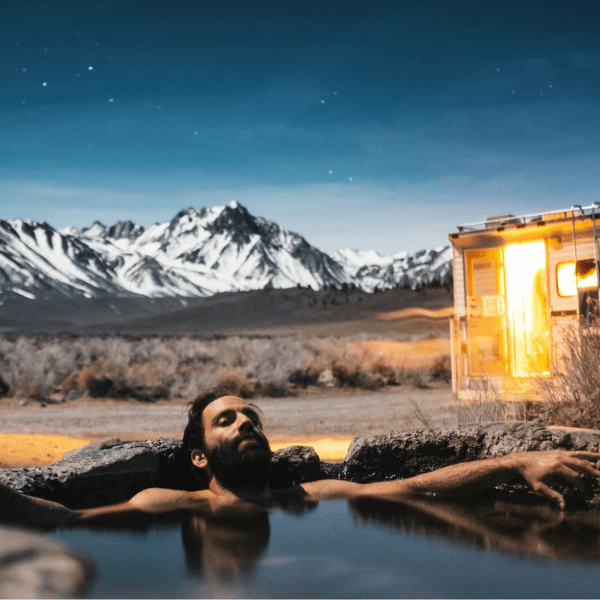 Man soaking in a natural hot spring at night with snow‑capped mountains in the background and a warmly lit RV parked nearby, representing eco‑responsible RV travel and a low‑impact way to enjoy the great outdoors.
