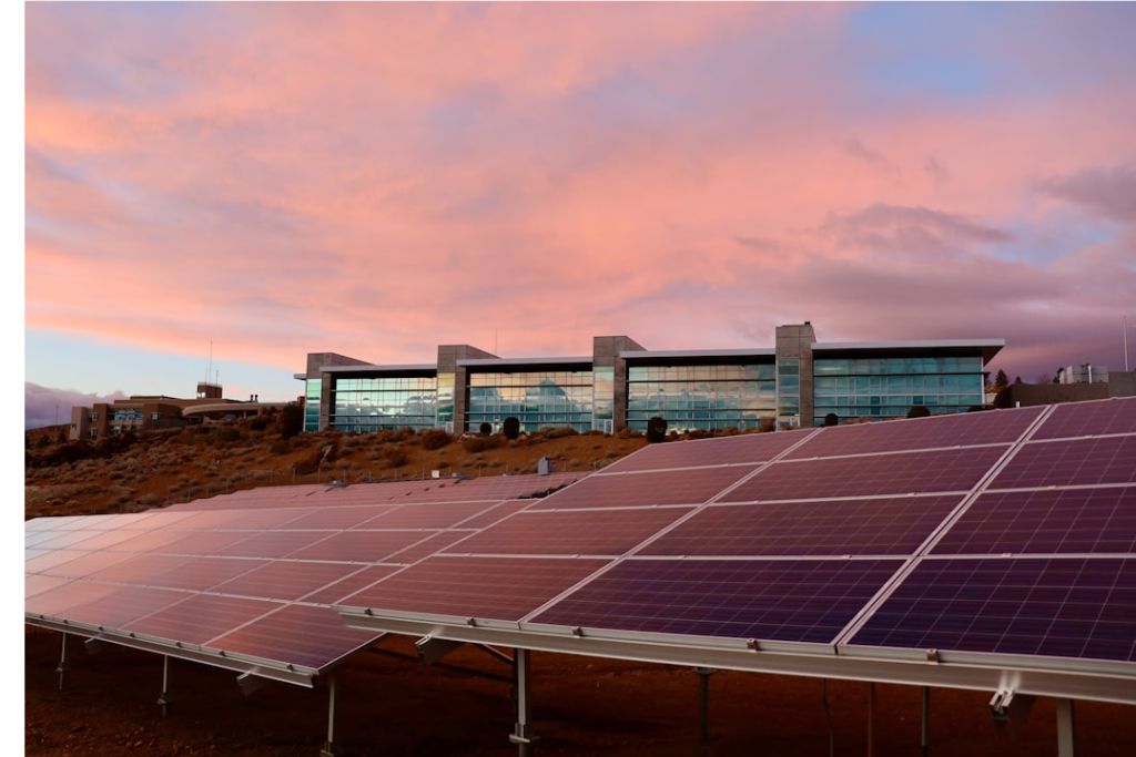Ground-mounted solar panels in front of a modern glass office building under a pink sunset sky