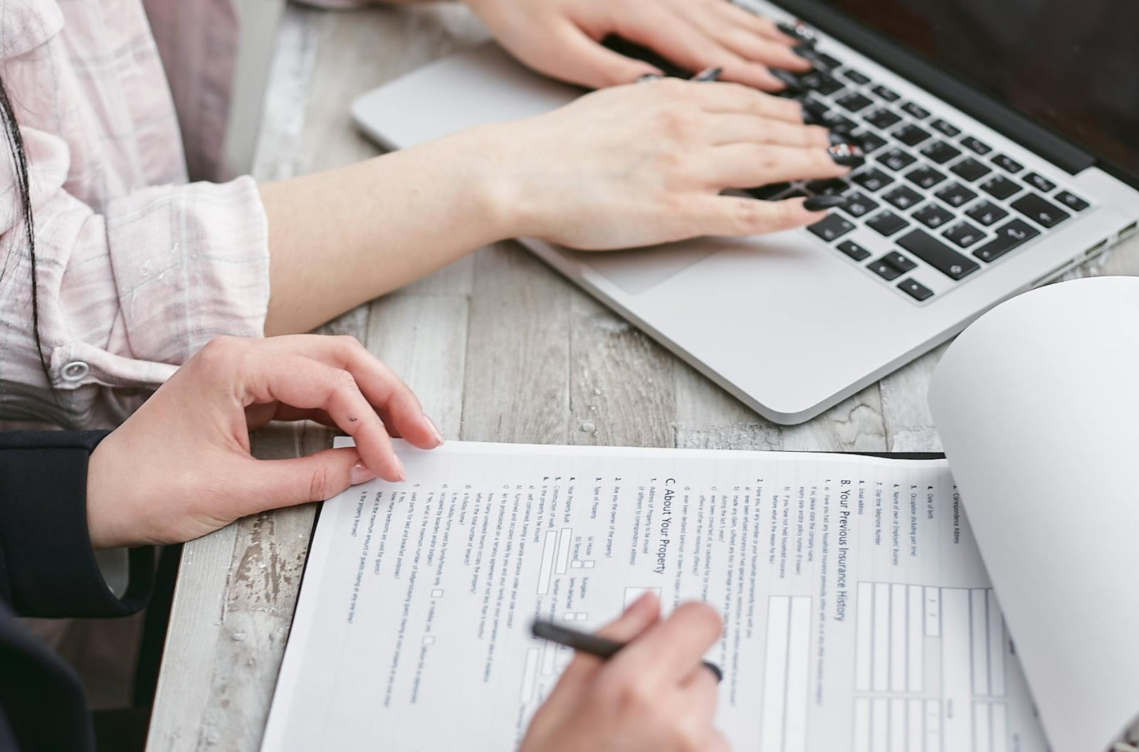 Two people working together at a wooden desk, one filling out a paper form with a pen while the other types on a laptop.