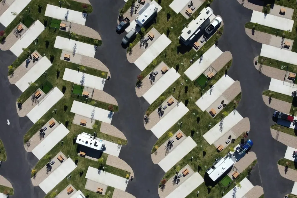 A top-down aerial view of a modern RV park with rows of angled concrete parking pads, each with a picnic table and small lawn, and several motorhomes and camper vans parked along dark asphalt roads.