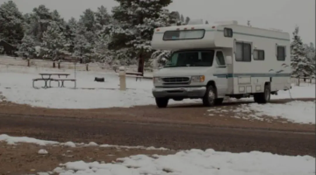 A white Ford E-350 Class C motorhome parked on a snowy dirt road at a forested campsite surrounded by snow-covered pine trees, a picnic table, and fire ring under a cloudy winter sky.