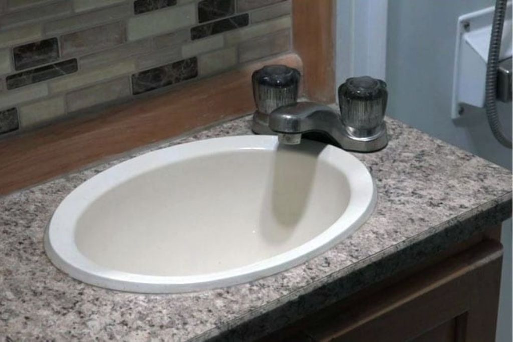 Oval bathroom sink installed in a speckled countertop with a dual-handle faucet and decorative tile backsplash in an RV